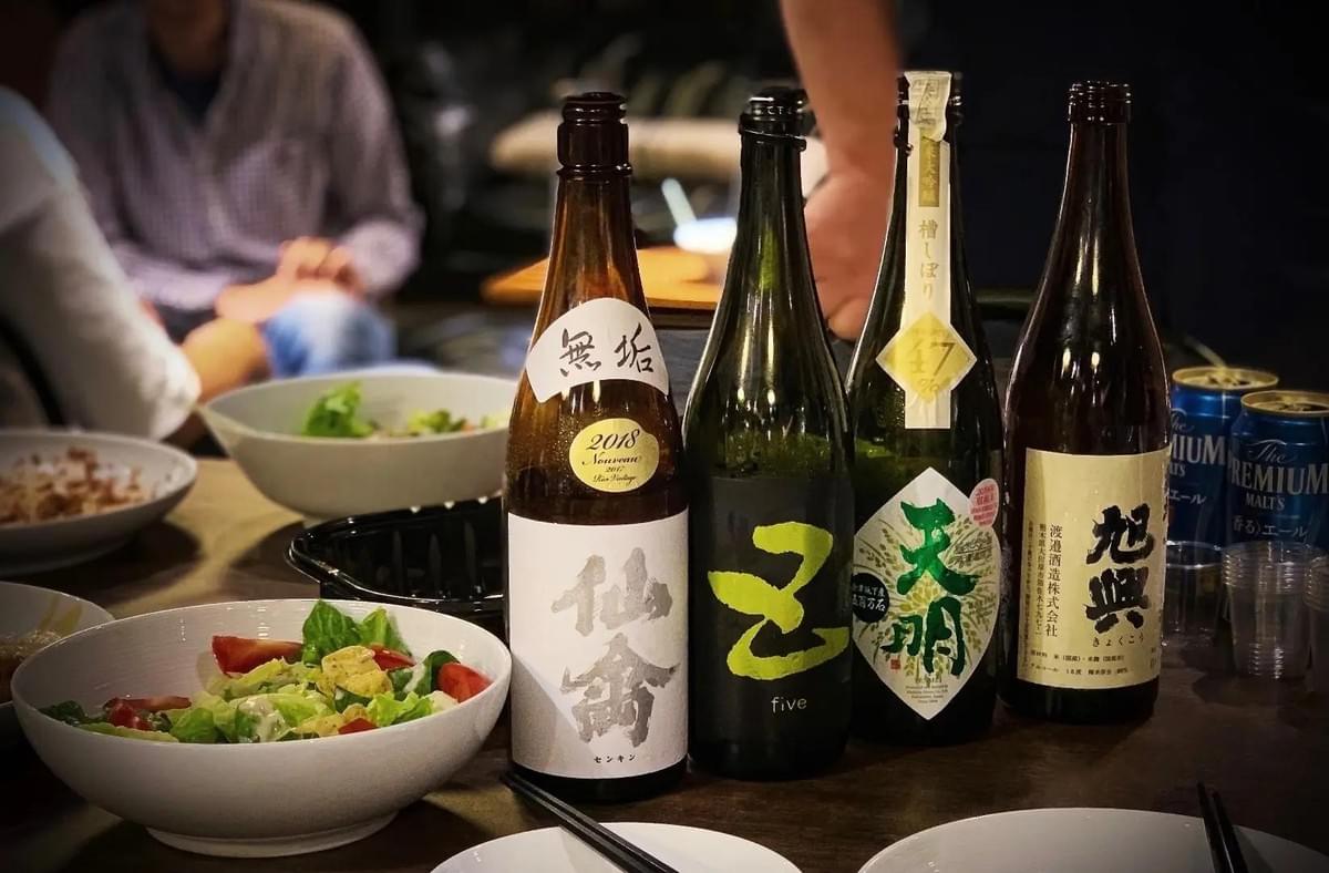 Four bottles of Japanese sake with distinct labels stand on a table alongside fresh salad bowls and blue cans of Suntory beer. The blurred background shows a cozy izakaya setting with people enjoying a meal in soft, warm lighting. Four bottles of Japanese sake with distinct labels stand on a table alongside fresh salad bowls and blue cans of Suntory beer. The blurred background shows a cozy izakaya setting with people enjoying a meal in soft, warm lighting.