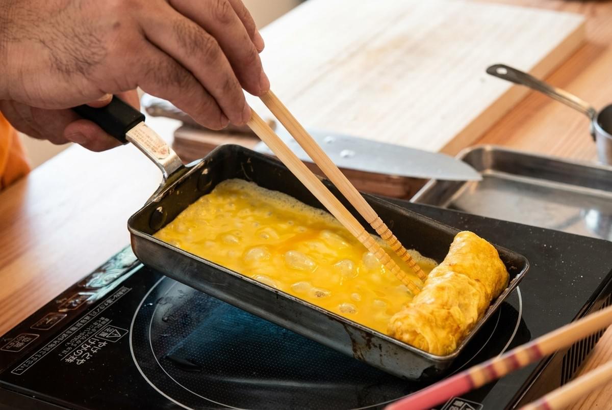 Cooking tamagoyaki by rolling liquid egg in a rectangular pan using long chopsticks. Cooking tamagoyaki by rolling liquid egg in a rectangular pan using long chopsticks.