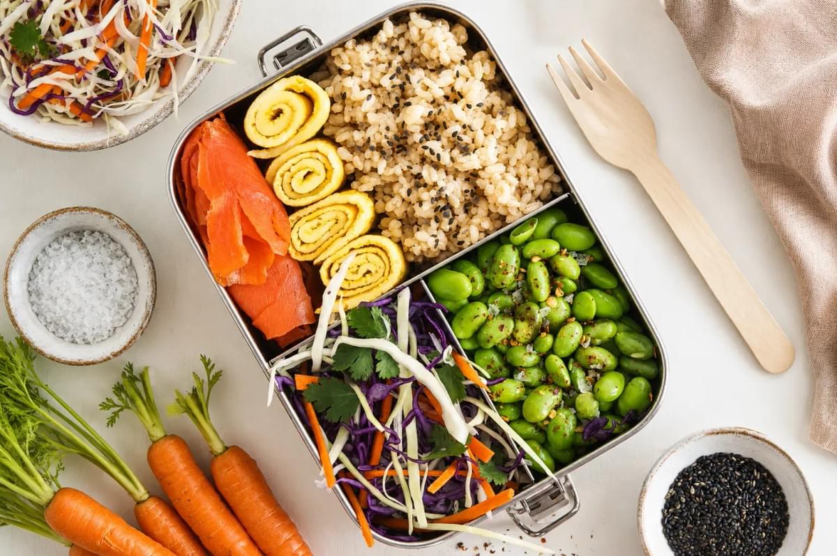 A stainless steel bento box packed with brown rice, sliced smoked salmon, tamagoyaki rolls, seasoned edamame, and a shredded cabbage salad. A stainless steel bento box packed with brown rice, sliced smoked salmon, tamagoyaki rolls, seasoned edamame, and a shredded cabbage salad.