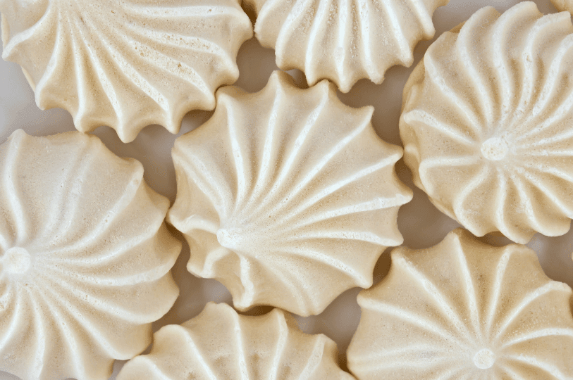 An overhead macro shot of several pale beige meringue cookies, focusing on the symmetrical, sunburst-like ridges and smooth matte finish of the baked egg whites. An overhead macro shot of several pale beige meringue cookies, focusing on the symmetrical, sunburst-like ridges and smooth matte finish of the baked egg whites.