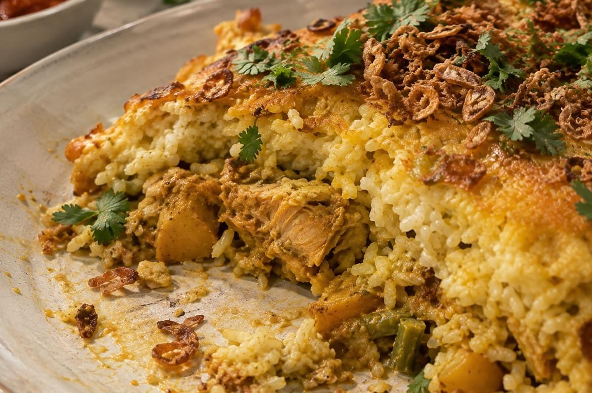 A close-up view of a slice taken from the curry chicken rice bake, detailing the moist, flavorful interior filled with chicken and potatoes underneath a baked, golden rice crust sprinkled with crispy shallots and fresh cilantro. A close-up view of a slice taken from the curry chicken rice bake, detailing the moist, flavorful interior filled with chicken and potatoes underneath a baked, golden rice crust sprinkled with crispy shallots and fresh cilantro.