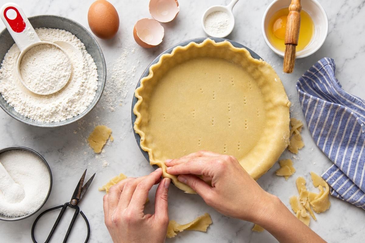 Top-down view of hands fluting the edge of raw pie dough in a baking pan alongside ingredients. Top-down view of hands fluting the edge of raw pie dough in a baking pan alongside ingredients.