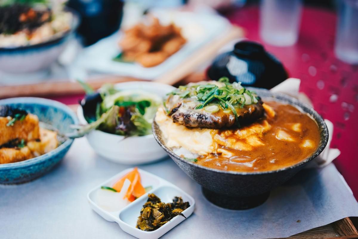 A close-up shot of a Japanese curry bowl featuring a hamburger patty topped with sliced green onions over a bed of egg and rice. The main dish is surrounded by various side dishes on a dining table, including a green salad, pickled vegetables, and tofu. A close-up shot of a Japanese curry bowl featuring a hamburger patty topped with sliced green onions over a bed of egg and rice. The main dish is surrounded by various side dishes on a dining table, including a green salad, pickled vegetables, and tofu.