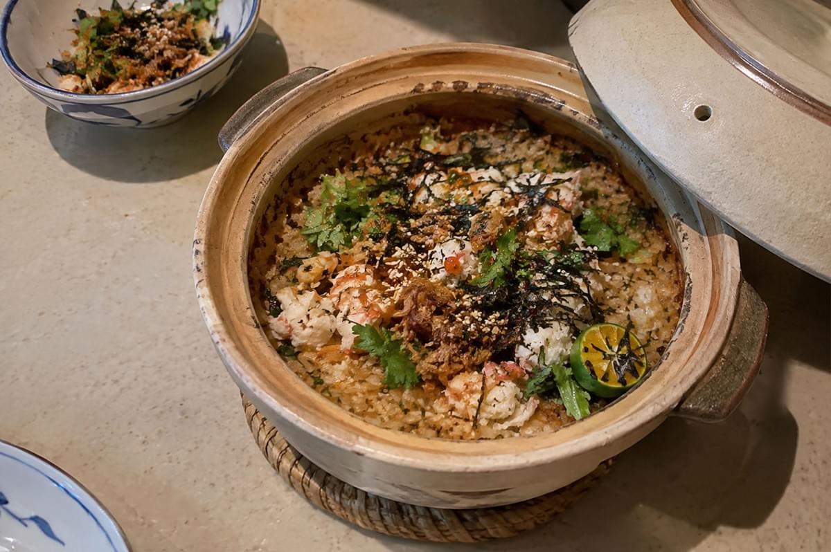 A high-angle view showcases a traditional Japanese ceramic donabe pot filled with seasoned rice topped with fresh crab meat, shredded nori seaweed, toasted sesame seeds, fresh cilantro leaves, and a grilled citrus half. The beige lid rests propped open on the right side of the pot, which sits on a woven trivet next to a small blue-and-white serving bowl filled with a portioned mix of the rice and toppings. A high-angle view showcases a traditional Japanese ceramic donabe pot filled with seasoned rice topped with fresh crab meat, shredded nori seaweed, toasted sesame seeds, fresh cilantro leaves, and a grilled citrus half. The beige lid rests propped open on the right side of the pot, which sits on a woven trivet next to a small blue-and-white serving bowl filled with a portioned mix of the rice and toppings.