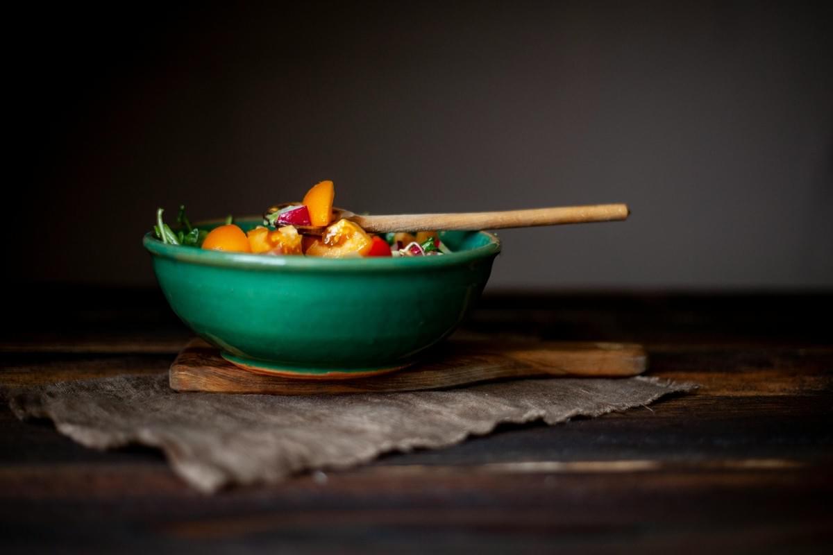 A vibrant salad filled with orange tomato wedges, radishes, and fresh greens sits in a bright turquoise ceramic bowl. The bowl rests on a rustic wooden cutting board and a coarse cloth napkin atop a dark wooden table against a moody, shadowed background. A vibrant salad filled with orange tomato wedges, radishes, and fresh greens sits in a bright turquoise ceramic bowl. The bowl rests on a rustic wooden cutting board and a coarse cloth napkin atop a dark wooden table against a moody, shadowed background.