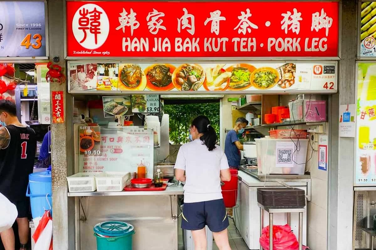 A woman stands at a Bak Kut Teh food stall with a bright red sign displaying dishes. The vendor prepares food in a compact, bustling environment. A woman stands at a Bak Kut Teh food stall with a bright red sign displaying dishes. The vendor prepares food in a compact, bustling environment.