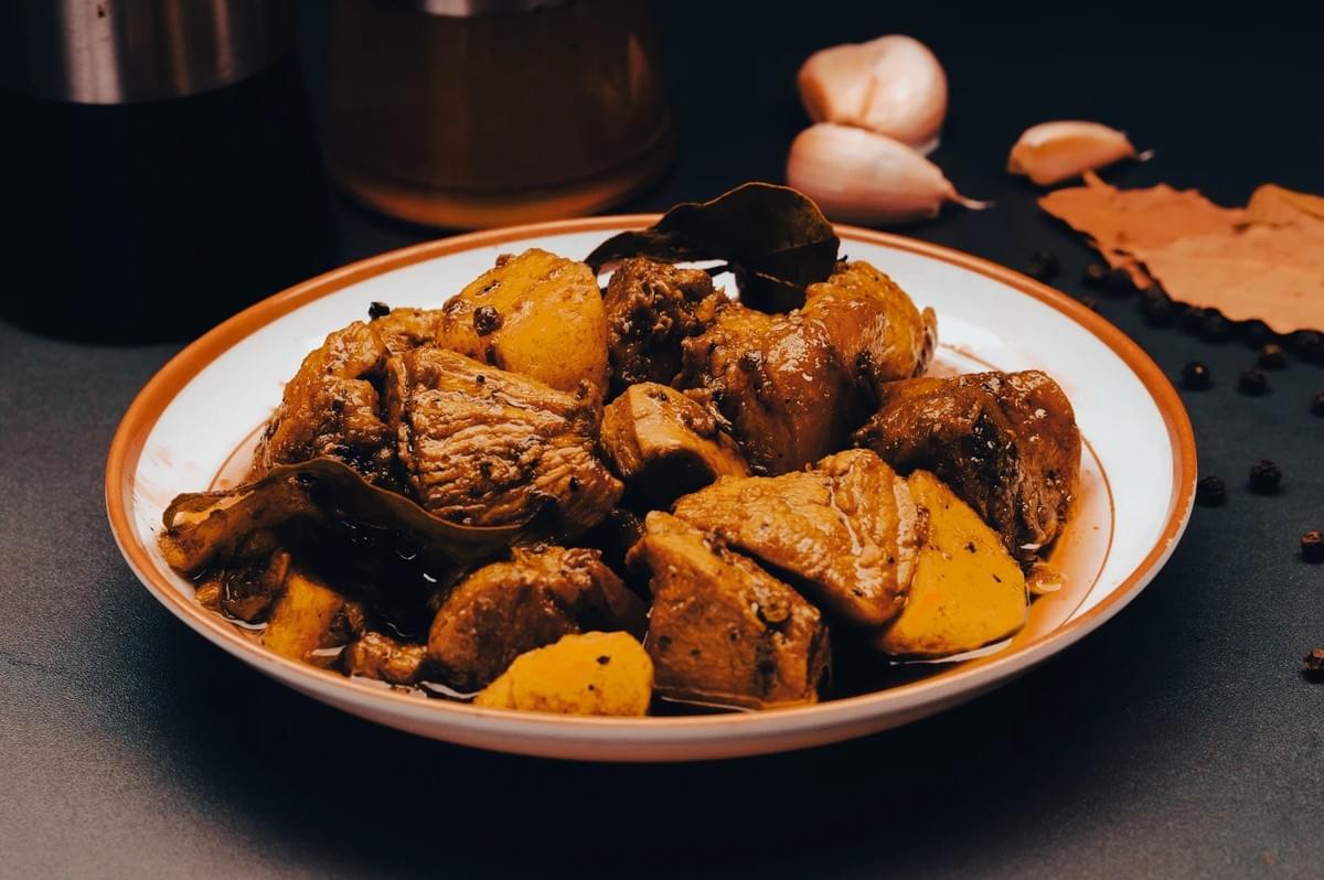 Close‑up of a plate of Filipino chicken adobo with tender chicken pieces, potatoes, bay leaves, and peppercorns in a rich soy‑vinegar sauce, surrounded by garlic and spices on a dark surface. Close‑up of a plate of Filipino chicken adobo with tender chicken pieces, potatoes, bay leaves, and peppercorns in a rich soy‑vinegar sauce, surrounded by garlic and spices on a dark surface.