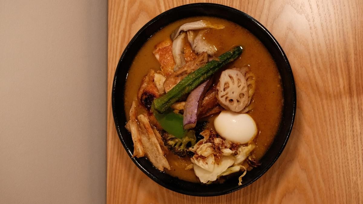 A round black bowl filled with savory soup curry rests on a light wooden table surface. The dish is topped with a soft-boiled egg, lotus root, okra, eggplant, mushrooms, and fried chicken. A round black bowl filled with savory soup curry rests on a light wooden table surface. The dish is topped with a soft-boiled egg, lotus root, okra, eggplant, mushrooms, and fried chicken.