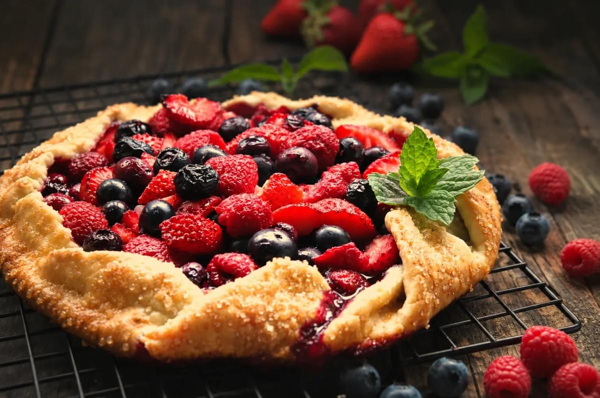 A close-up, angled shot of a freshly baked rustic berry galette cooling on a black wire rack. The golden-brown crust is sprinkled with coarse sugar and filled with a vibrant mix of strawberries, raspberries, and blueberries, topped with a fresh sprig of mint. Extra loose berries and mint leaves scatter across the dark wooden table background. A close-up, angled shot of a freshly baked rustic berry galette cooling on a black wire rack. The golden-brown crust is sprinkled with coarse sugar and filled with a vibrant mix of strawberries, raspberries, and blueberries, topped with a fresh sprig of mint. Extra loose berries and mint leaves scatter across the dark wooden table background.