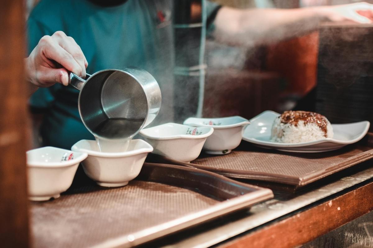 A person's hand is shown pouring steaming liquid from a metal ladle into small white bowls arranged on a tray. In the background, a plate of white rice topped with a dark sauce sits ready for service at a food stall. A person's hand is shown pouring steaming liquid from a metal ladle into small white bowls arranged on a tray. In the background, a plate of white rice topped with a dark sauce sits ready for service at a food stall.