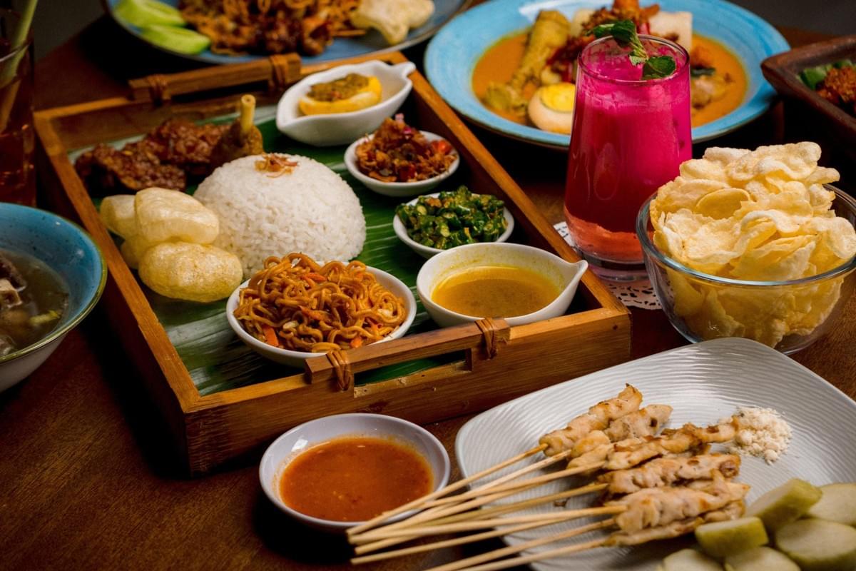 A vibrant spread of traditional Indonesian food is arranged on a wooden table, featuring a central wooden platter with white rice, fried noodles, skewers, crackers, and various side dishes. Surrounding the tray are plates of chicken satay with dipping sauce, a bowl of curry, a glass of bright pink dragon fruit juice, and a bowl of crispy chips. A vibrant spread of traditional Indonesian food is arranged on a wooden table, featuring a central wooden platter with white rice, fried noodles, skewers, crackers, and various side dishes. Surrounding the tray are plates of chicken satay with dipping sauce, a bowl of curry, a glass of bright pink dragon fruit juice, and a bowl of crispy chips.