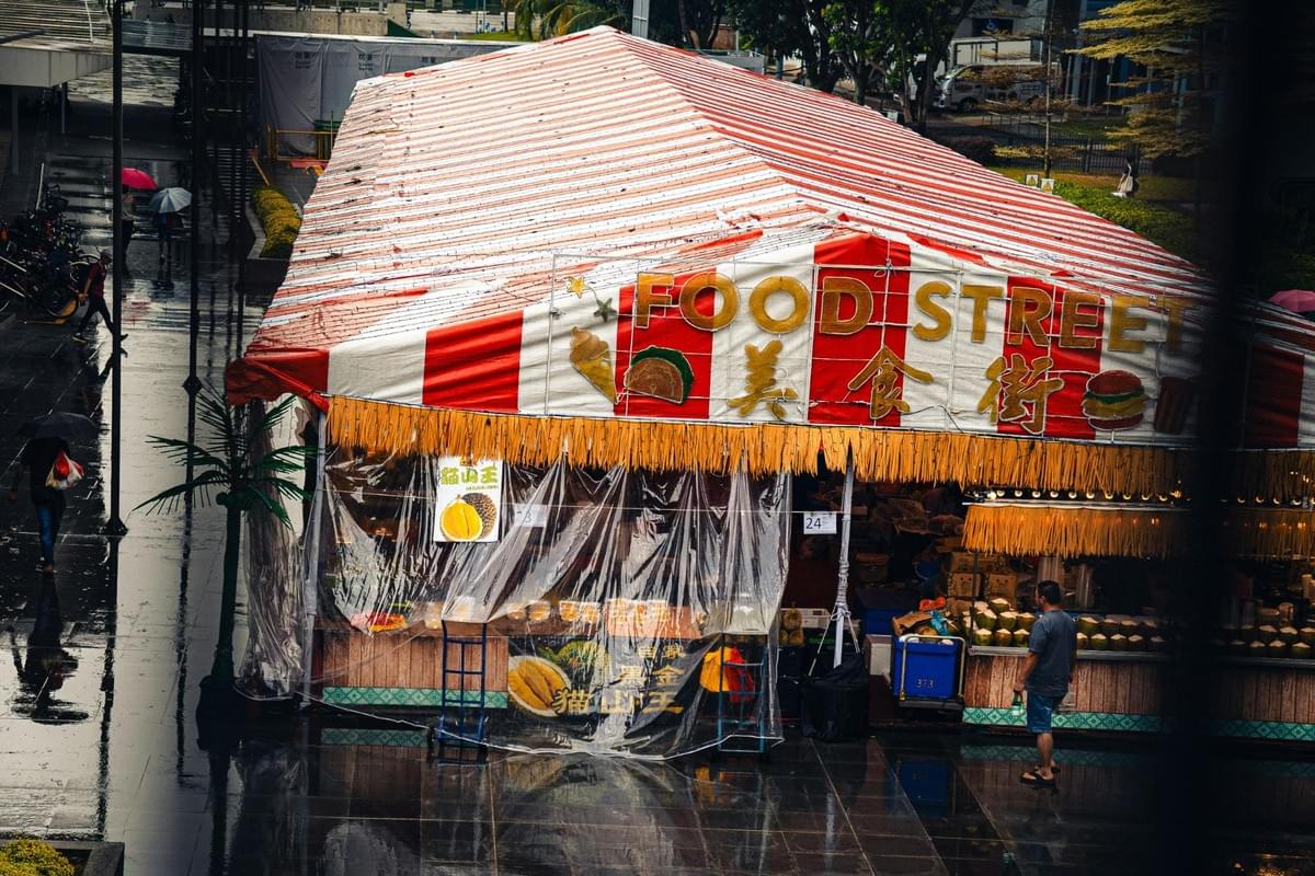 A large, red-and-white striped tent labeled "FOOD STREET" stands prominently on a wet, reflective pavement during a rainy day. Passersby with umbrellas navigate the area near the stall, which features gold lettering and displays of various food items like durian and coconuts. A large, red-and-white striped tent labeled "FOOD STREET" stands prominently on a wet, reflective pavement during a rainy day. Passersby with umbrellas navigate the area near the stall, which features gold lettering and displays of various food items like durian and coconuts.