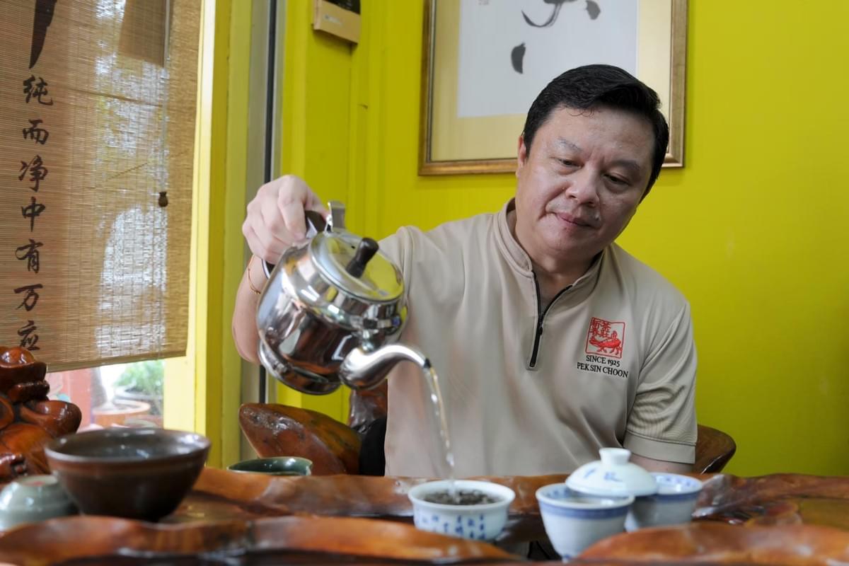 A man carefully pours hot water from a silver kettle into a small ceramic teacup set on a wooden tea tray. He wears a beige shirt featuring the Pek Sin Choon logo inside a room with vibrant yellow walls and framed Chinese artwork. A man carefully pours hot water from a silver kettle into a small ceramic teacup set on a wooden tea tray. He wears a beige shirt featuring the Pek Sin Choon logo inside a room with vibrant yellow walls and framed Chinese artwork.