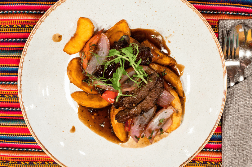 An overhead gourmet presentation of Lomo Saltado on a white ceramic plate. Thick-cut potato wedges and strips of seared sirloin are drizzled with a rich brown sauce and topped with delicate microgreens, set against a colorful Peruvian textile background. An overhead gourmet presentation of Lomo Saltado on a white ceramic plate. Thick-cut potato wedges and strips of seared sirloin are drizzled with a rich brown sauce and topped with delicate microgreens, set against a colorful Peruvian textile background.