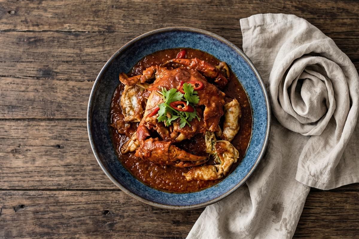 Overhead view of a plated crab dish coated in rich red sauce and garnished with sliced chilies and fresh cilantro on a blue ceramic plate. Overhead view of a plated crab dish coated in rich red sauce and garnished with sliced chilies and fresh cilantro on a blue ceramic plate.
