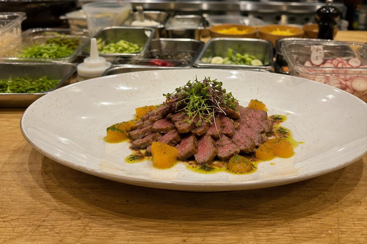 Showcases a beautifully plated, gourmet dish set against the backdrop of a professional prep station. At the center of a large, shallow white bowl, thin slices of medium-rare beef are neatly fanned out in a circular arrangement, topped with a delicate pile of fresh microgreens. The meat is garnished with vibrant orange segments and drizzled with a herb-infused oil and dark reduction. In the blurred background, various stainless steel containers and small wooden bowls hold organized ingredients like sliced radishes, chopped greens, and sauces, highlighting the precision and creative process of a culinary environment. Showcases a beautifully plated, gourmet dish set against the backdrop of a professional prep station. At the center of a large, shallow white bowl, thin slices of medium-rare beef are neatly fanned out in a circular arrangement, topped with a delicate pile of fresh microgreens. The meat is garnished with vibrant orange segments and drizzled with a herb-infused oil and dark reduction. In the blurred background, various stainless steel containers and small wooden bowls hold organized ingredients like sliced radishes, chopped greens, and sauces, highlighting the precision and creative process of a culinary environment.