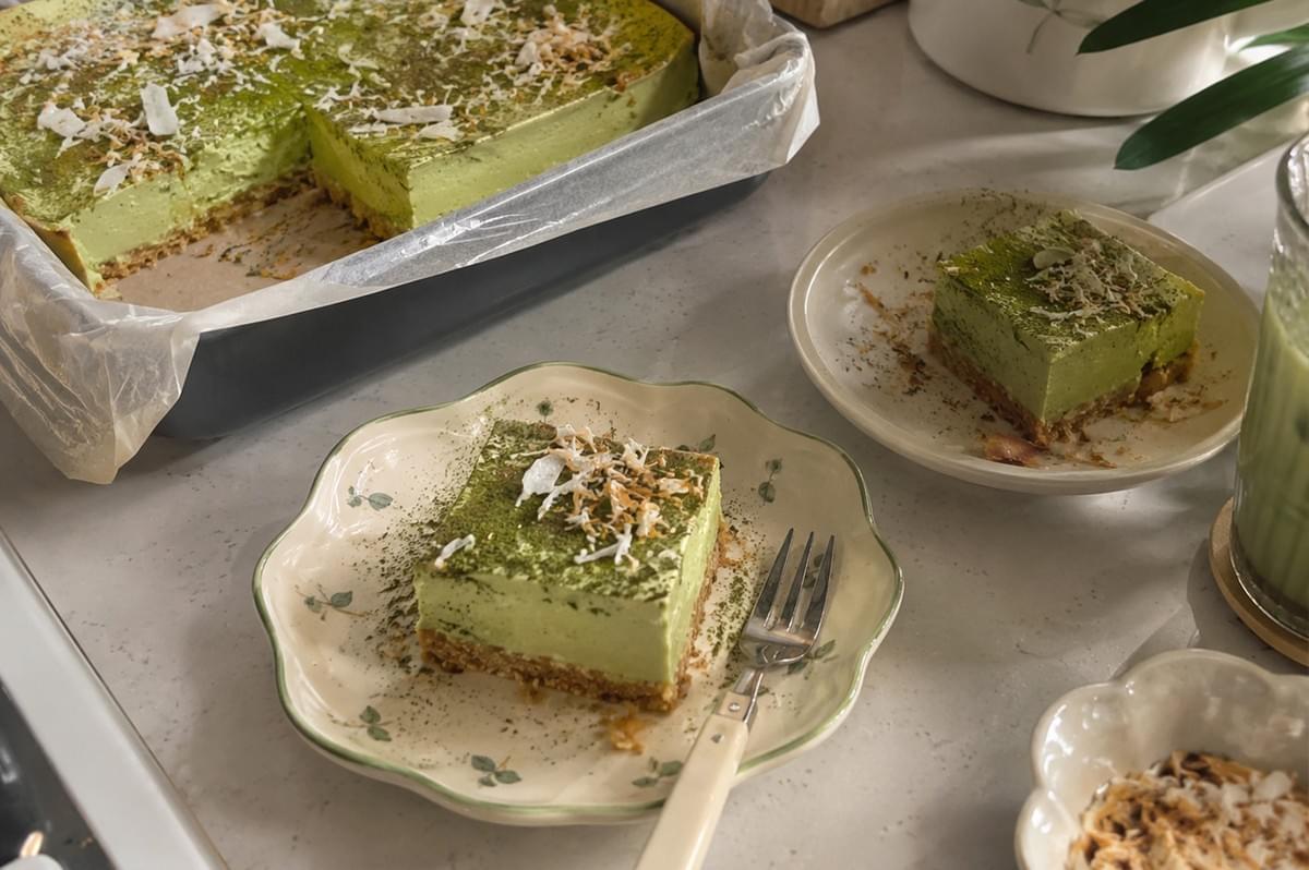 An overhead shot displays a freshly baked batch of light-green pandan matcha cheesecake sitting inside a parchment-lined square baking pan with a corner slice removed. In the foreground, a single square cheesecake bar topped with matcha dusting and toasted coconut flakes rests on a scalloped floral ceramic plate next to a fork, with another plated bar, a dish of toasted coconut, and an iced matcha drink arranged nearby on the marble countertop. An overhead shot displays a freshly baked batch of light-green pandan matcha cheesecake sitting inside a parchment-lined square baking pan with a corner slice removed. In the foreground, a single square cheesecake bar topped with matcha dusting and toasted coconut flakes rests on a scalloped floral ceramic plate next to a fork, with another plated bar, a dish of toasted coconut, and an iced matcha drink arranged nearby on the marble countertop.