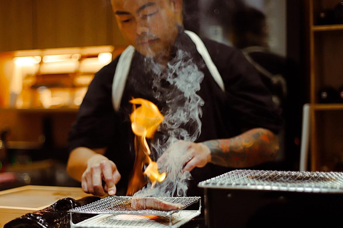 Chef grilling meat over an open flame at an omakase counter, with smoke rising from the yakiniku-style grill. Chef grilling meat over an open flame at an omakase counter, with smoke rising from the yakiniku-style grill.