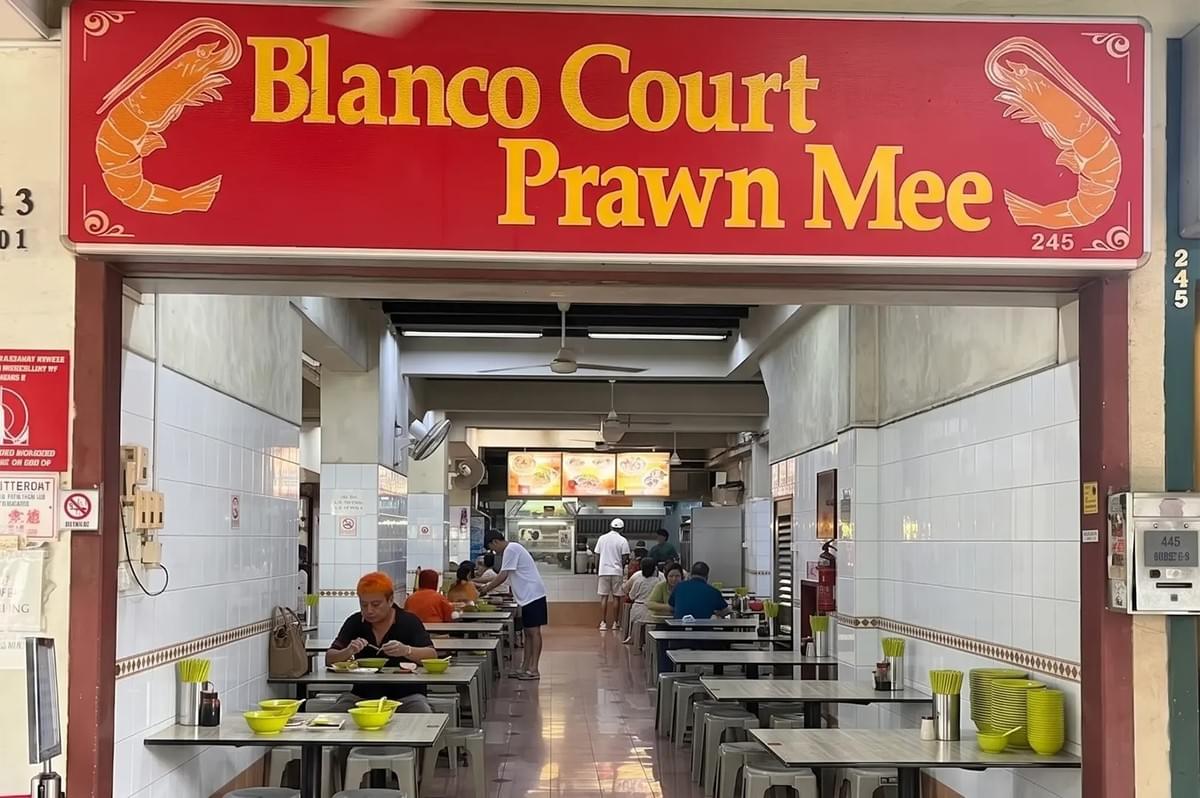 Entrance of Blanco Court Prawn Mee in Singapore with diners at indoor tables under a red sign. Entrance of Blanco Court Prawn Mee in Singapore with diners at indoor tables under a red sign.