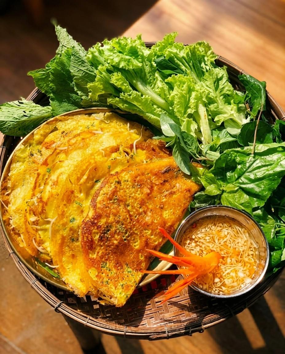 A high-angle shot of Vietnamese bánh xèo—crispy, golden-yellow savory crepes—served in a woven basket alongside a mountain of fresh green lettuce, herbs, and a small bowl of dipping sauce garnished with a carved carrot flower. A high-angle shot of Vietnamese bánh xèo—crispy, golden-yellow savory crepes—served in a woven basket alongside a mountain of fresh green lettuce, herbs, and a small bowl of dipping sauce garnished with a carved carrot flower.