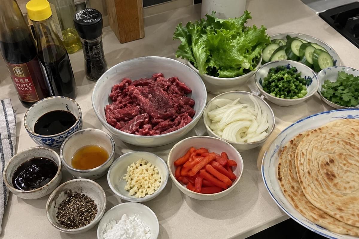 A high-angle prep-shot featuring an array of raw ingredients arranged in small ceramic bowls on a kitchen counter for making black pepper beef roti wraps. At the center is a large bowl of sliced raw beef, surrounded by bowls of fresh lettuce, sliced green onions, cilantro, cucumbers, red bell peppers, sliced white onions, minced garlic, cornstarch, and coarse ground black pepper. A stack of cooked, golden-brown roti flatbreads rests on a plate on the right, while bottles of oyster sauce and soy sauce stand on the left. A high-angle prep-shot featuring an array of raw ingredients arranged in small ceramic bowls on a kitchen counter for making black pepper beef roti wraps. At the center is a large bowl of sliced raw beef, surrounded by bowls of fresh lettuce, sliced green onions, cilantro, cucumbers, red bell peppers, sliced white onions, minced garlic, cornstarch, and coarse ground black pepper. A stack of cooked, golden-brown roti flatbreads rests on a plate on the right, while bottles of oyster sauce and soy sauce stand on the left.