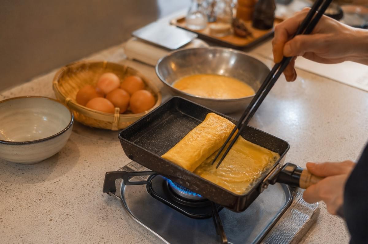 A cook uses long black chopsticks to lift and fold a thin layer of cooking egg mixture over an already rolled omelet inside a rectangular, non-stick tamagoyaki pan resting on a gas stove. A woven basket of fresh brown eggs and a bowl of whisked egg mixture sit on the counter in the background. A cook uses long black chopsticks to lift and fold a thin layer of cooking egg mixture over an already rolled omelet inside a rectangular, non-stick tamagoyaki pan resting on a gas stove. A woven basket of fresh brown eggs and a bowl of whisked egg mixture sit on the counter in the background.