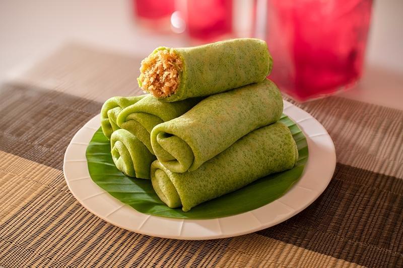 A stack of green pandan crepes, filled with grated coconut and rolled up, sits on a banana leaf on a white plate. One rolled crepe on top is sliced open, revealing the sweet, brown palm sugar-infused coconut filling inside. A stack of green pandan crepes, filled with grated coconut and rolled up, sits on a banana leaf on a white plate. One rolled crepe on top is sliced open, revealing the sweet, brown palm sugar-infused coconut filling inside.