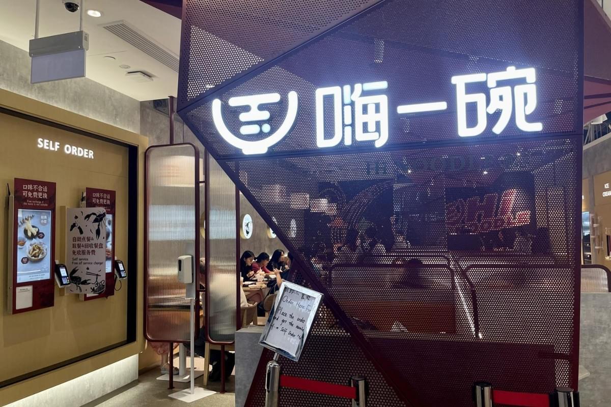 An illuminated sign featuring Chinese characters and a bowl logo marks the entrance of a modern noodle restaurant enclosed in dark red mesh paneling. To the left, digital self-ordering kiosks are mounted on the wall near a partitioned dining area with seated customers. An illuminated sign featuring Chinese characters and a bowl logo marks the entrance of a modern noodle restaurant enclosed in dark red mesh paneling. To the left, digital self-ordering kiosks are mounted on the wall near a partitioned dining area with seated customers.