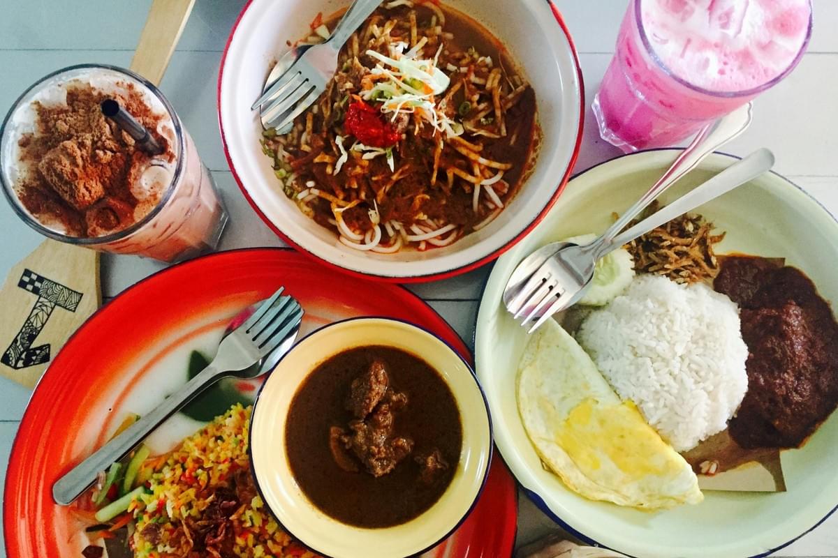The image shows a top-down view of a colorful Malaysian meal featuring plates of nasi lemak with an egg, fried rice with a side of beef curry, and a noodle dish. Accompanying the food are two vibrant drinks, including a pink iced beverage and a chocolate malt drink topped with cocoa powder. The image shows a top-down view of a colorful Malaysian meal featuring plates of nasi lemak with an egg, fried rice with a side of beef curry, and a noodle dish. Accompanying the food are two vibrant drinks, including a pink iced beverage and a chocolate malt drink topped with cocoa powder.