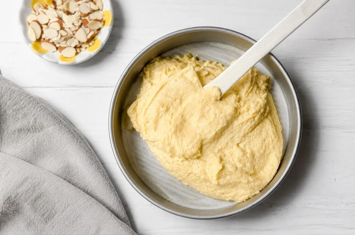 Thick cake batter spread in a round baking pan next to a small dish of sliced almonds. Thick cake batter spread in a round baking pan next to a small dish of sliced almonds.