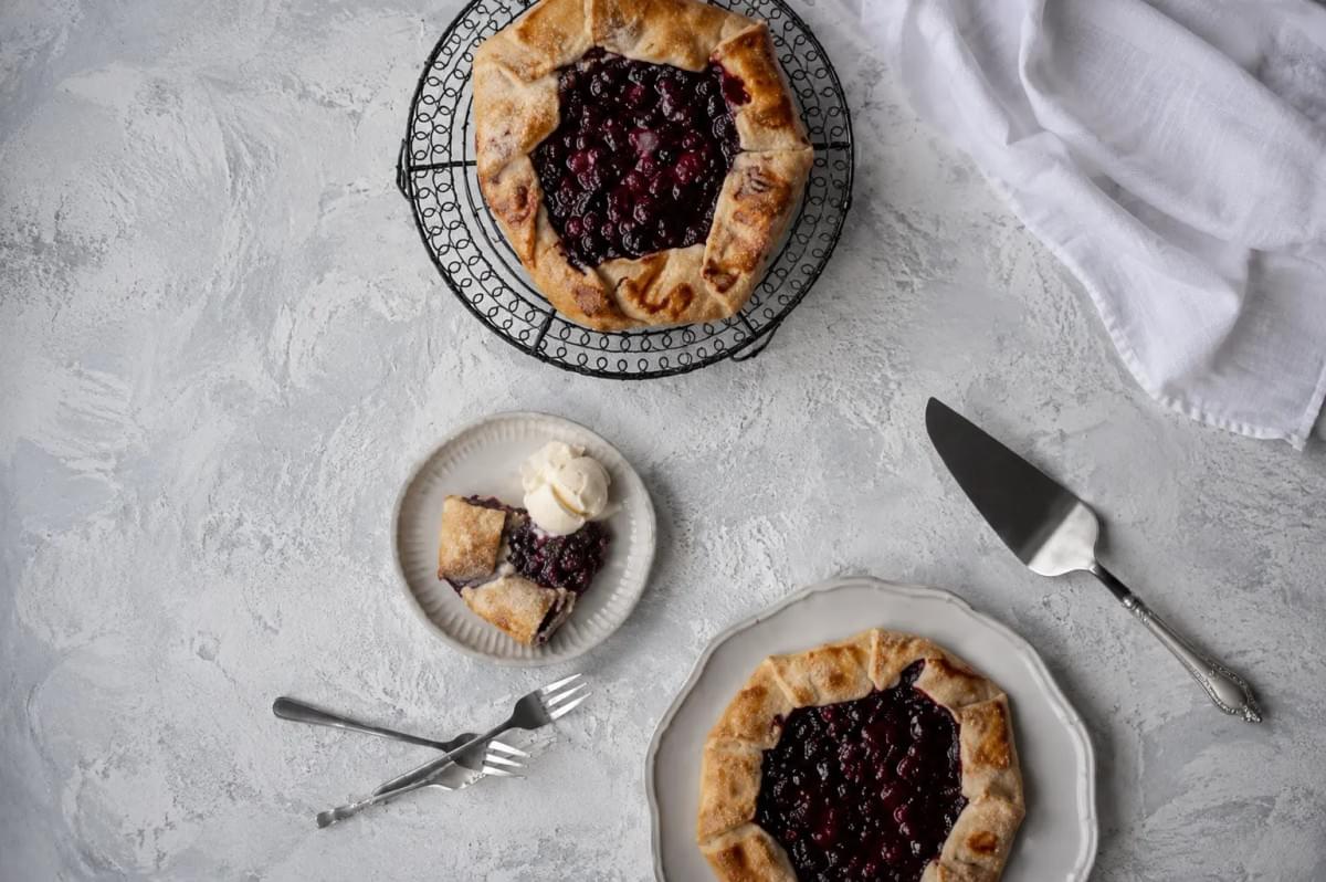 The arrangement includes two whole berry galettes with golden-brown, pleated crusts—one resting on a black wire cooling rack and the other on a ceramic plate. A small slice of the galette is served on a separate saucer alongside a scoop of vanilla ice cream, accompanied by two dessert forks, a pie server, and a crumpled white cloth. The arrangement includes two whole berry galettes with golden-brown, pleated crusts—one resting on a black wire cooling rack and the other on a ceramic plate. A small slice of the galette is served on a separate saucer alongside a scoop of vanilla ice cream, accompanied by two dessert forks, a pie server, and a crumpled white cloth.