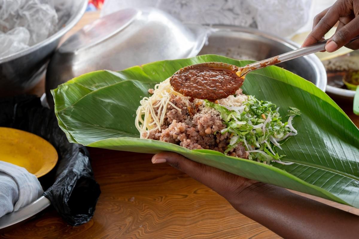 A vendor holds a wide green leaf supporting a dish made of rice, noodles, and fresh greens. Another person uses a metal spoon to pour a savory, dark sauce directly over the meal. A vendor holds a wide green leaf supporting a dish made of rice, noodles, and fresh greens. Another person uses a metal spoon to pour a savory, dark sauce directly over the meal.