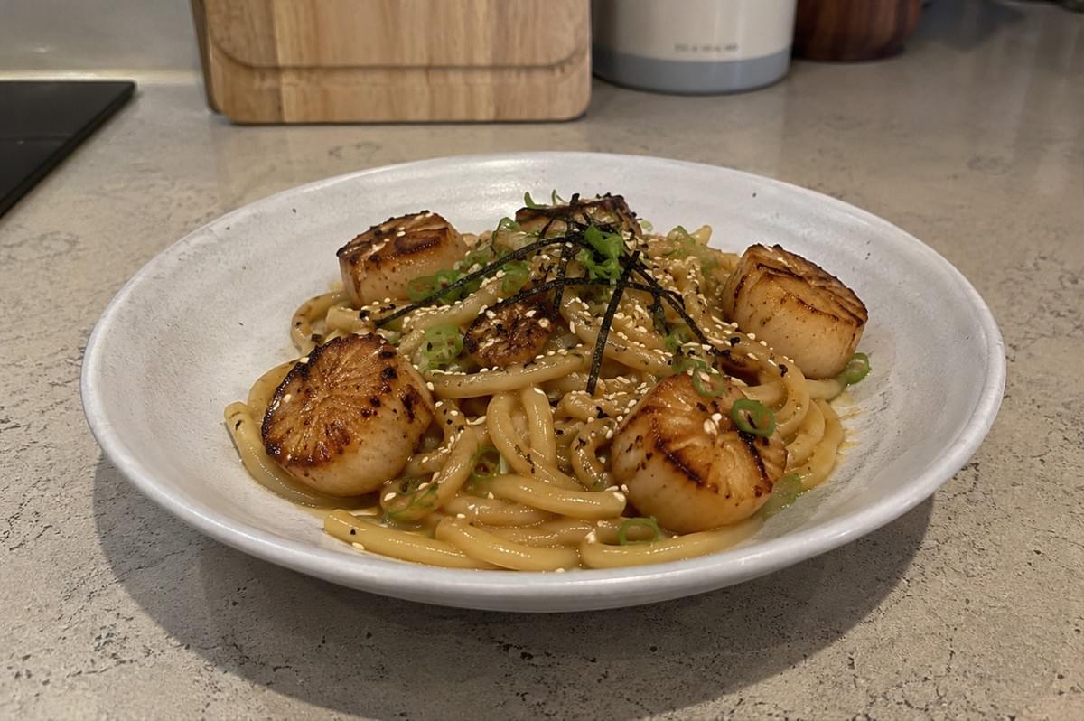 Features a shallow, light-grey speckled ceramic bowl filled with thick udon noodles coated in a glossy, light brown miso butter sauce. Five large, plump sea scallops with a deep golden-brown sear on top are arranged over the noodles. The dish is beautifully garnished with a sprinkle of white sesame seeds, finely chopped green onions, and thin strips of black nori (seaweed) clustered in the center. The bowl rests on a neutral, speckled beige countertop with a soft-focus kitchen background showing a wooden cutting board and kitchen canisters. Features a shallow, light-grey speckled ceramic bowl filled with thick udon noodles coated in a glossy, light brown miso butter sauce. Five large, plump sea scallops with a deep golden-brown sear on top are arranged over the noodles. The dish is beautifully garnished with a sprinkle of white sesame seeds, finely chopped green onions, and thin strips of black nori (seaweed) clustered in the center. The bowl rests on a neutral, speckled beige countertop with a soft-focus kitchen background showing a wooden cutting board and kitchen canisters.