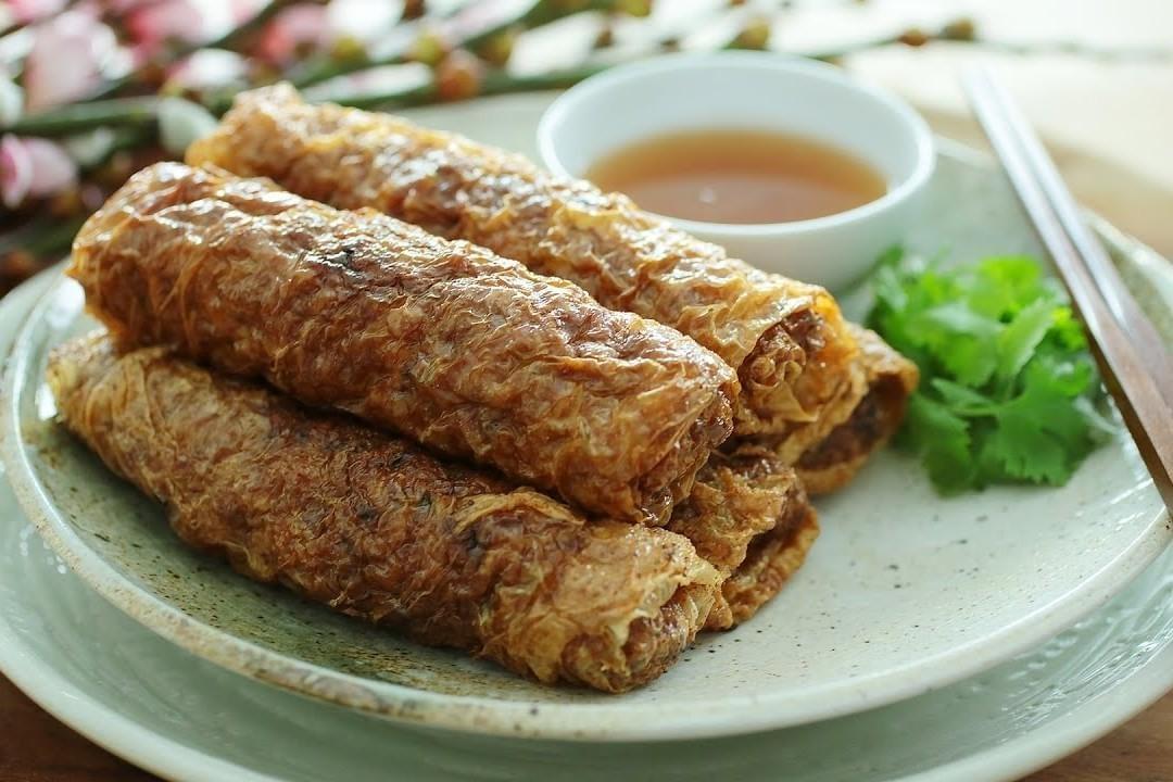 A stack of golden-brown, crispy fried tofu skin rolls arranged on a light-colored ceramic plate. The plate is garnished with fresh cilantro sprigs and served alongside a small white bowl of dipping sauce and a pair of chopsticks. A stack of golden-brown, crispy fried tofu skin rolls arranged on a light-colored ceramic plate. The plate is garnished with fresh cilantro sprigs and served alongside a small white bowl of dipping sauce and a pair of chopsticks.
