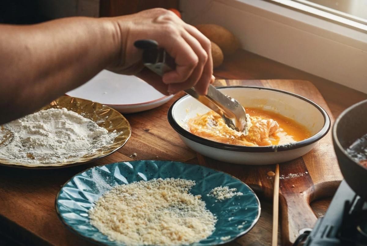 Person dipping raw chicken into egg wash at a breading station with flour and panko. Person dipping raw chicken into egg wash at a breading station with flour and panko.