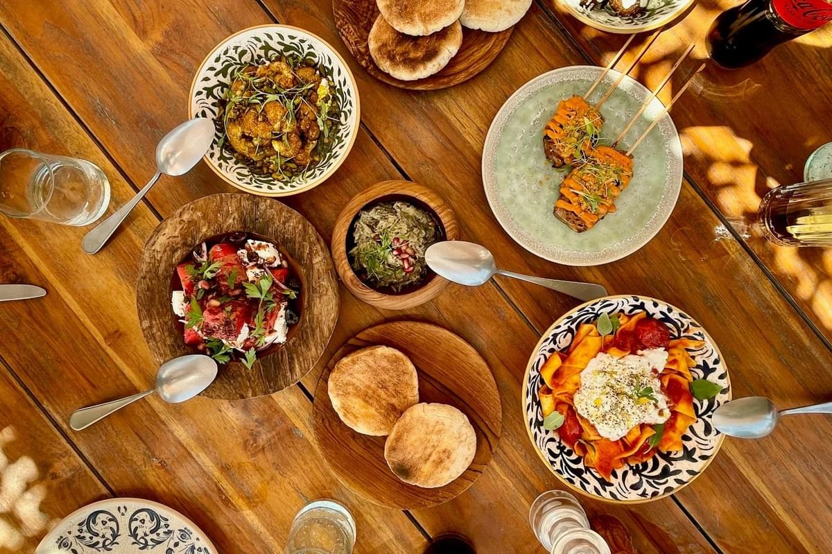 A rustic wooden table is abundantly set with a variety of Mediterranean-inspired dishes, including fresh salads, pita bread, pasta, and skewers. Dappled sunlight casts patterned shadows across the spread, creating a warm and inviting atmosphere for a shared meal. A rustic wooden table is abundantly set with a variety of Mediterranean-inspired dishes, including fresh salads, pita bread, pasta, and skewers. Dappled sunlight casts patterned shadows across the spread, creating a warm and inviting atmosphere for a shared meal.