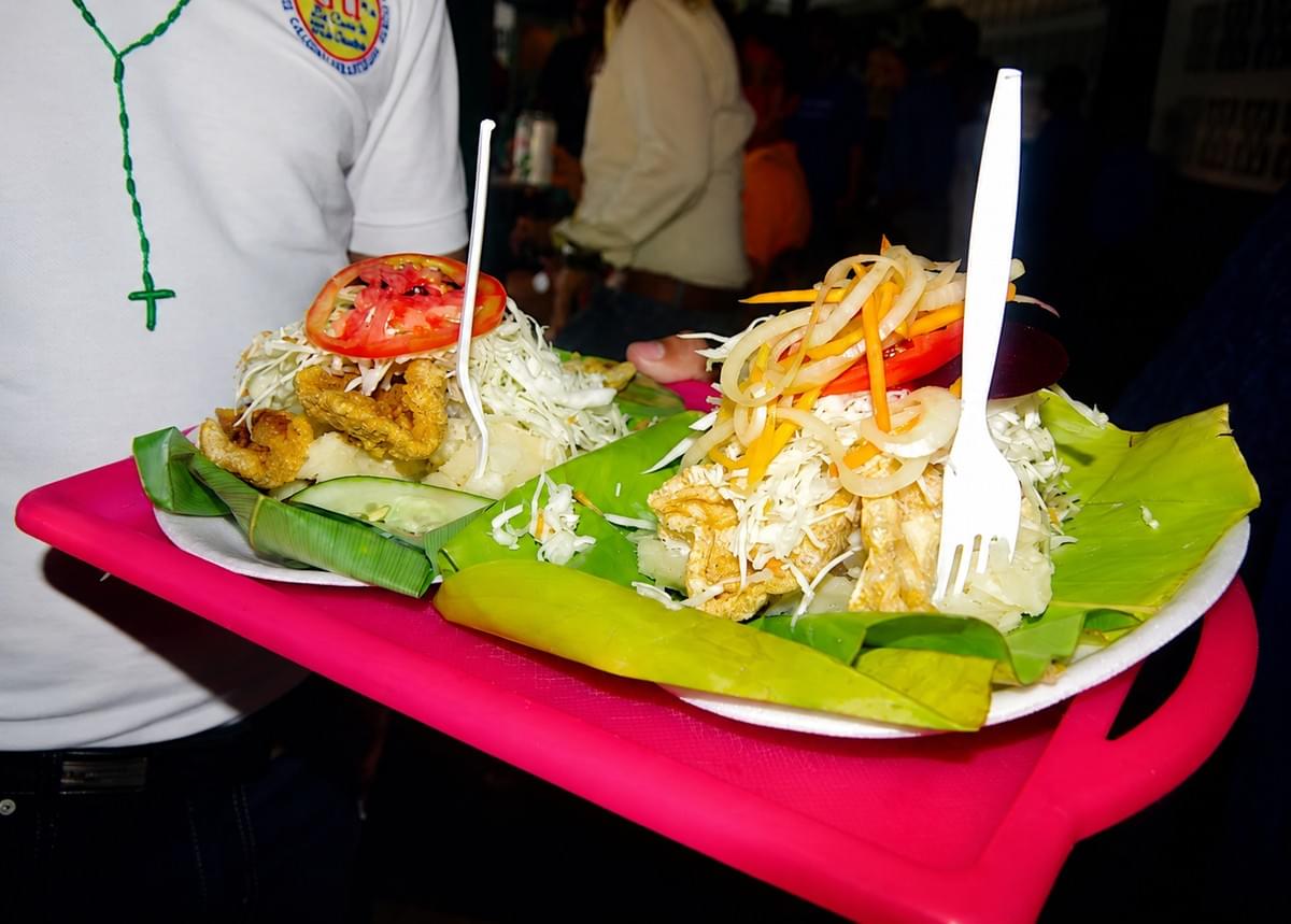 Carried on an eye-catching hot pink plastic tray, this lively street-festival shot bursts with electric energy, contrasting bright green banana leaves and ruby-red tomato slices against the busy background of a local crowd. Carried on an eye-catching hot pink plastic tray, this lively street-festival shot bursts with electric energy, contrasting bright green banana leaves and ruby-red tomato slices against the busy background of a local crowd.