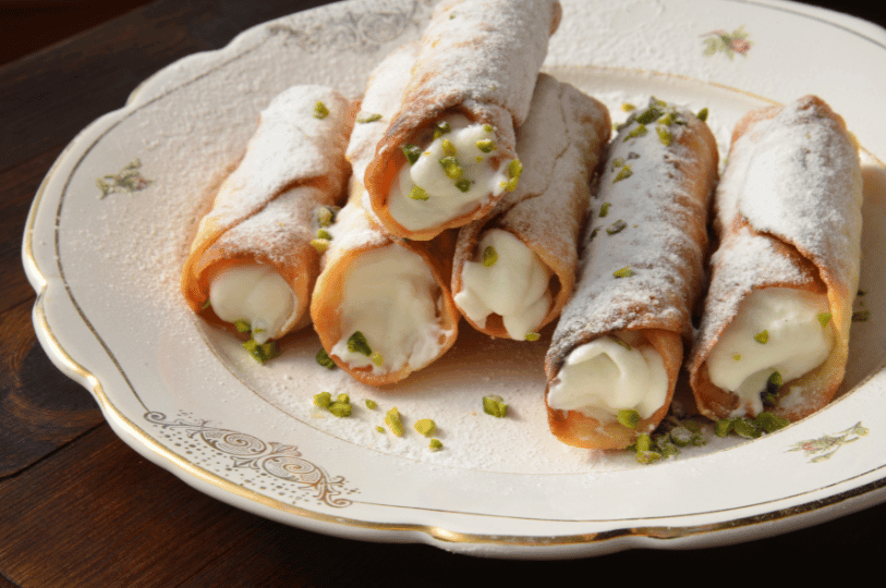 A sophisticated presentation of five cannoli stacked on a vintage floral-patterned china plate, garnished with a sprinkle of bright green pistachio bits and a fine layer of powdered sugar. A sophisticated presentation of five cannoli stacked on a vintage floral-patterned china plate, garnished with a sprinkle of bright green pistachio bits and a fine layer of powdered sugar.