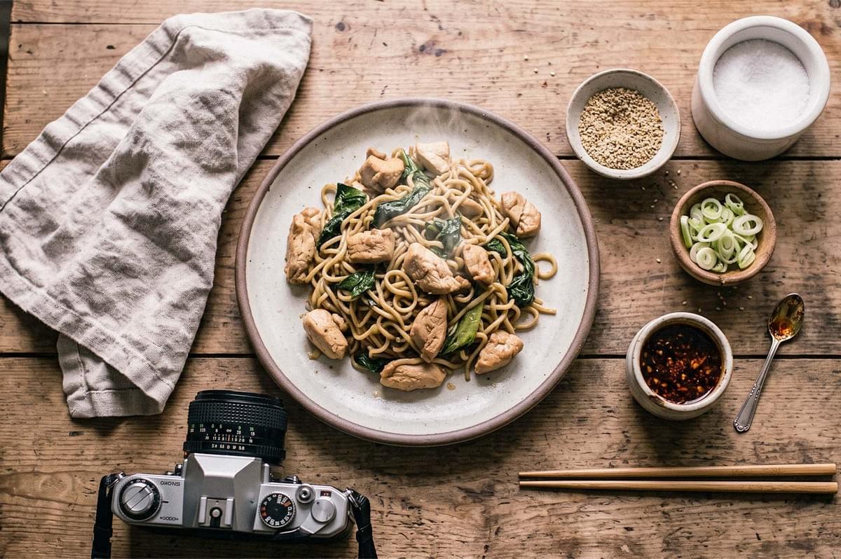Plate of chicken noodles with leafy greens on a rustic wooden table, surrounded by sesame seeds, sliced scallions, chili sauce, chopsticks, and a camera. Plate of chicken noodles with leafy greens on a rustic wooden table, surrounded by sesame seeds, sliced scallions, chili sauce, chopsticks, and a camera.