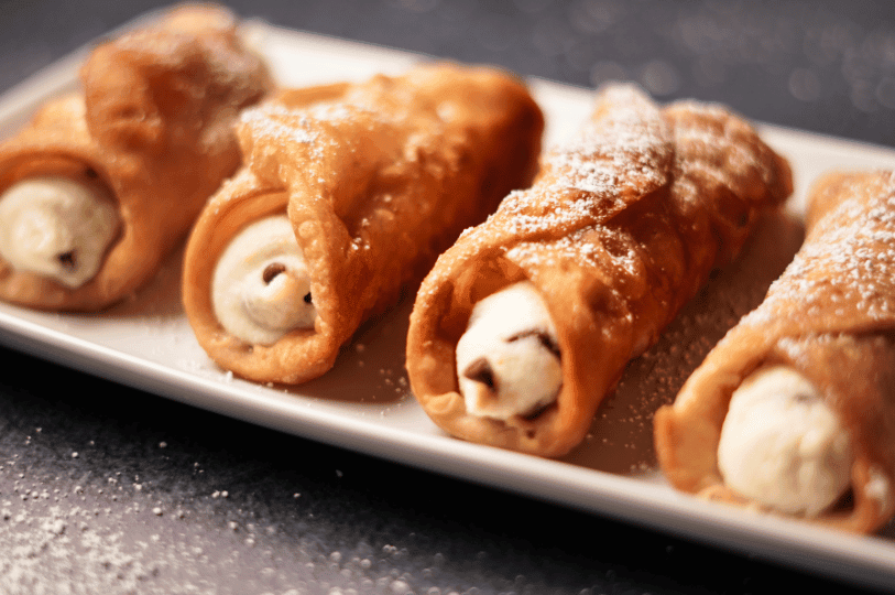 Four golden-brown, crispy cannoli shells lined up on a white rectangular plate, filled with smooth ricotta cream and lightly dusted with powdered sugar. Four golden-brown, crispy cannoli shells lined up on a white rectangular plate, filled with smooth ricotta cream and lightly dusted with powdered sugar.