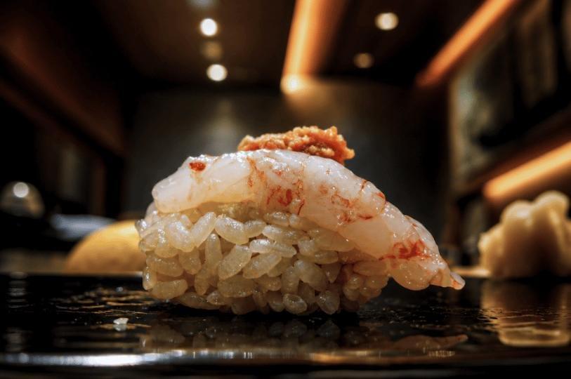 A close-up shot of a single piece of nigiri sushi featuring translucent raw sweet shrimp over seasoned rice, topped with a small garnish. The sushi sits on a glossy black plate in a dimly lit, warm restaurant setting. A close-up shot of a single piece of nigiri sushi featuring translucent raw sweet shrimp over seasoned rice, topped with a small garnish. The sushi sits on a glossy black plate in a dimly lit, warm restaurant setting.
