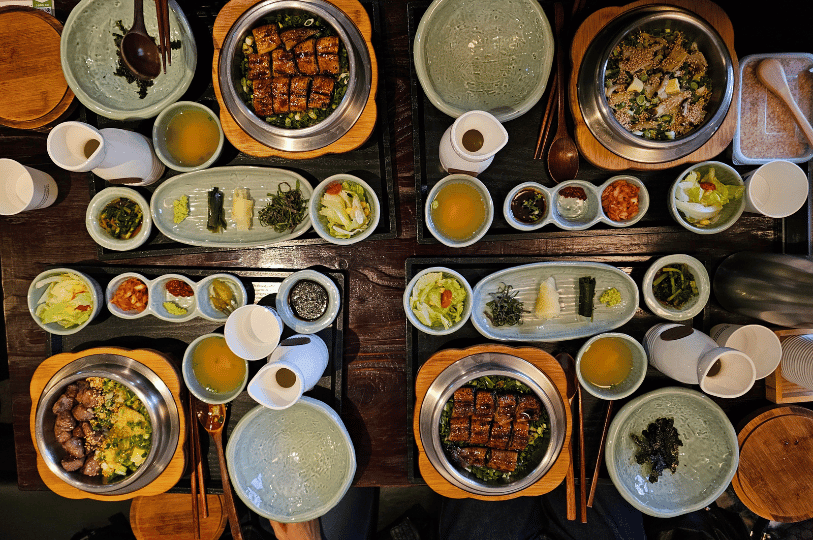 An elegant overhead shot of four individual Korean table settings on a dark wooden surface. Each setting features a main dish in a hot stone bowl, harmoniously accompanied by small, compartmentalized dishes of colorful banchan and individual soups. An elegant overhead shot of four individual Korean table settings on a dark wooden surface. Each setting features a main dish in a hot stone bowl, harmoniously accompanied by small, compartmentalized dishes of colorful banchan and individual soups.