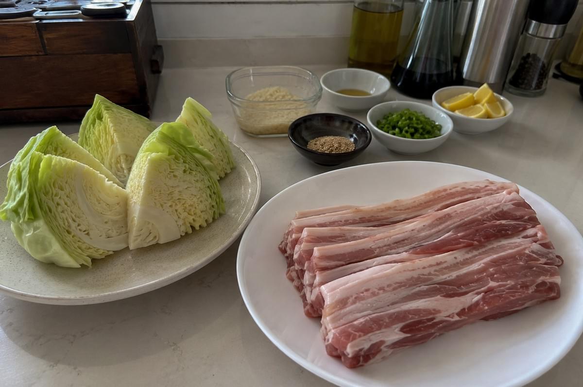 A bright, top-down kitchen counter shot showcasing the prepared raw ingredients for a meal arranged neatly on a light countertop. On the left, a ceramic plate holds four large, raw wedges of pale green cabbage, while a white plate on the right contains a neat stack of sliced, raw pork belly strips showing distinct layers of meat and fat. In the background, several small condiment bowls are spread out, containing sesame seeds, chopped green scallions, lemon wedges, panko breadcrumbs, and liquid ingredients alongside cooking oil and pepper grinders. A bright, top-down kitchen counter shot showcasing the prepared raw ingredients for a meal arranged neatly on a light countertop. On the left, a ceramic plate holds four large, raw wedges of pale green cabbage, while a white plate on the right contains a neat stack of sliced, raw pork belly strips showing distinct layers of meat and fat. In the background, several small condiment bowls are spread out, containing sesame seeds, chopped green scallions, lemon wedges, panko breadcrumbs, and liquid ingredients alongside cooking oil and pepper grinders.