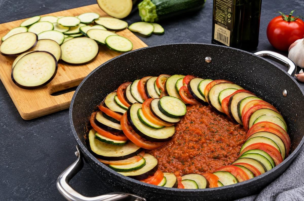 Slices of eggplant, zucchini, and tomato being arranged in a ring over tomato sauce in a pan, next to extra sliced vegetables on a wooden cutting board. Slices of eggplant, zucchini, and tomato being arranged in a ring over tomato sauce in a pan, next to extra sliced vegetables on a wooden cutting board.