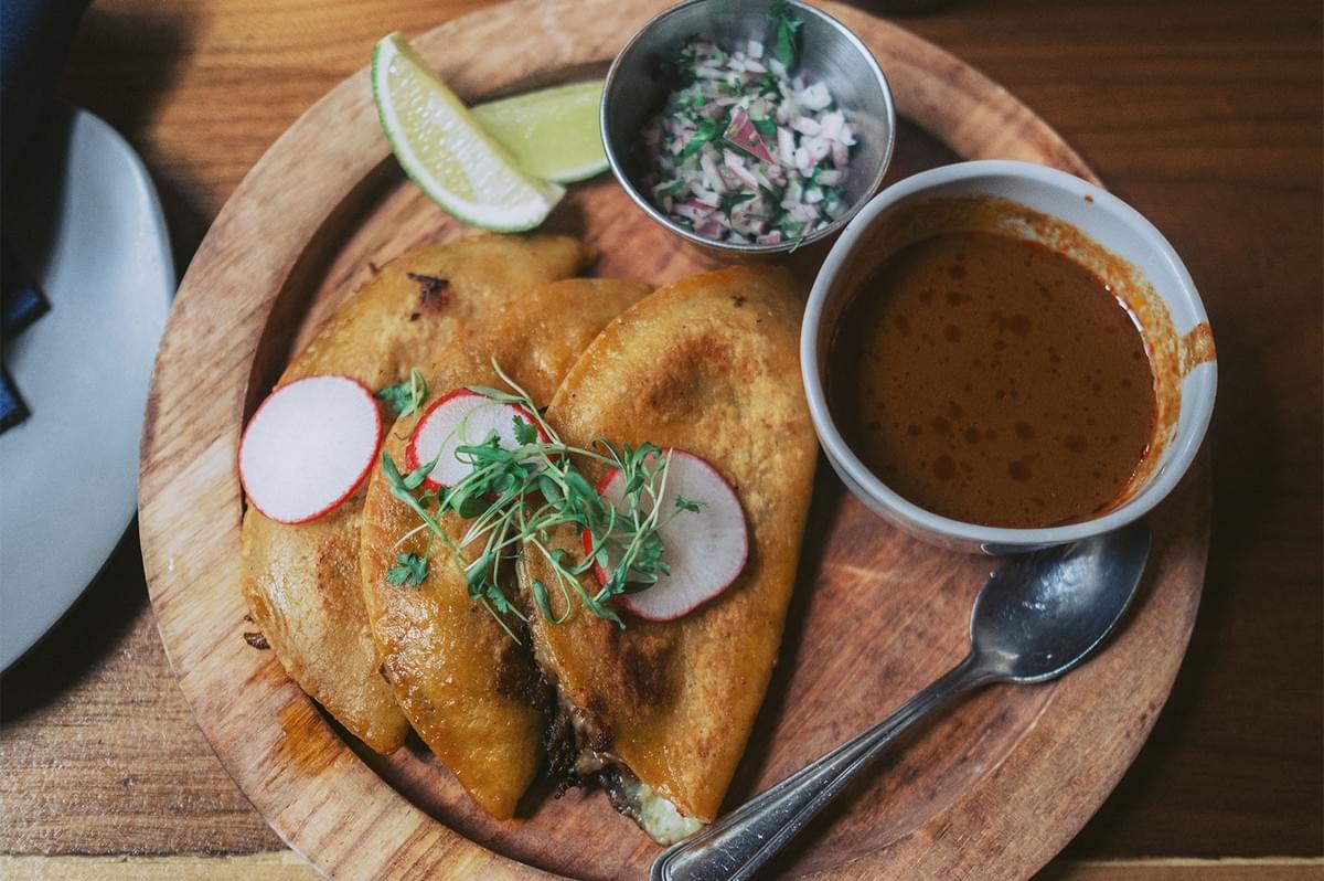 Three crispy birria tacos garnished with fresh radish and cilantro served on a wooden plate with lime, salsa, and consommé. Three crispy birria tacos garnished with fresh radish and cilantro served on a wooden plate with lime, salsa, and consommé.