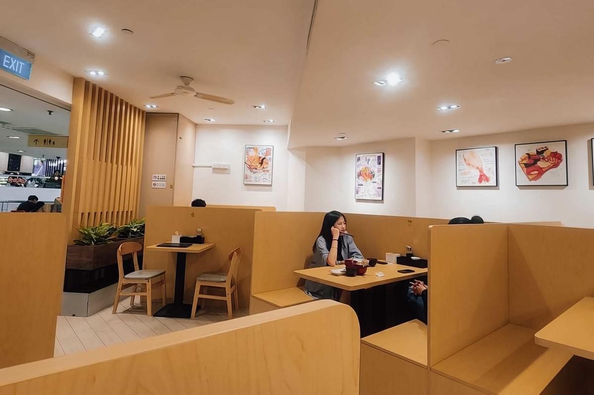 This wide-angle interior view captures the dining area of Chirashizushi Shou, featuring rows of high-backed light-wood booth seating under bright ceiling recessed lights. A customer sits at one of the central wooden tables, while framed culinary artwork hangs along the cream-colored back walls and vertical wooden slats screen off the left entrance area. This wide-angle interior view captures the dining area of Chirashizushi Shou, featuring rows of high-backed light-wood booth seating under bright ceiling recessed lights. A customer sits at one of the central wooden tables, while framed culinary artwork hangs along the cream-colored back walls and vertical wooden slats screen off the left entrance area.