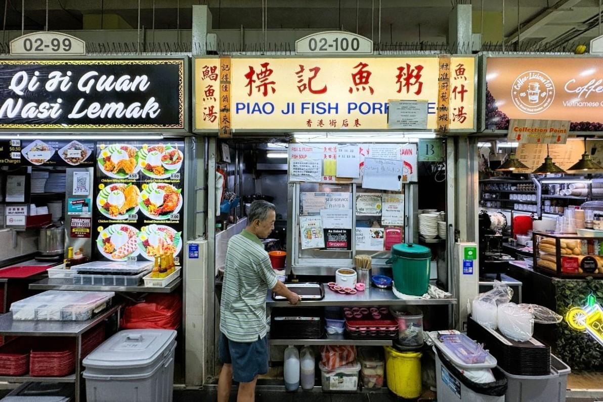 A hawker stall named "Piao Ji Fish Porridge" features yellow signage with blue and red lettering in a food center. A man in a striped shirt stands at the counter preparing orders between adjacent food stalls. A hawker stall named "Piao Ji Fish Porridge" features yellow signage with blue and red lettering in a food center. A man in a striped shirt stands at the counter preparing orders between adjacent food stalls.