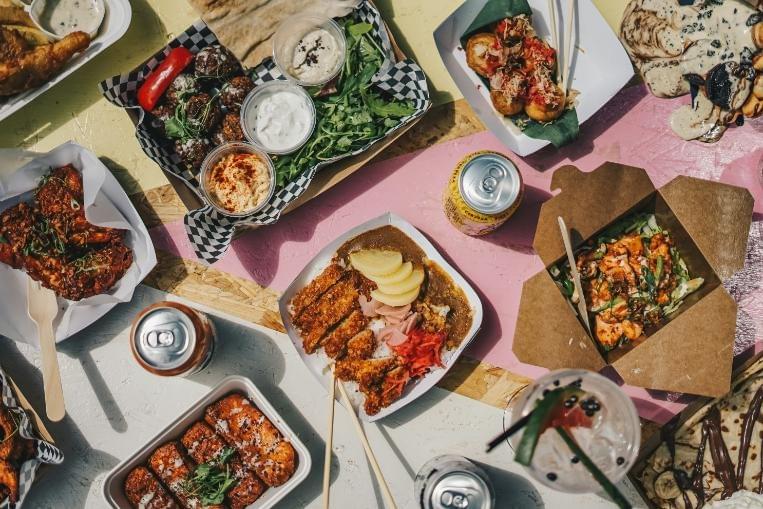 An overhead, top-down shot of the image displays a vibrant street food feast spread across a colorful pink and white background. The diverse spread includes takeout containers of Japanese chicken katsu curry, fried chicken wings, falafel with dips, takoyaki, and sweet desserts, alongside various canned and iced beverages. An overhead, top-down shot of the image displays a vibrant street food feast spread across a colorful pink and white background. The diverse spread includes takeout containers of Japanese chicken katsu curry, fried chicken wings, falafel with dips, takoyaki, and sweet desserts, alongside various canned and iced beverages.