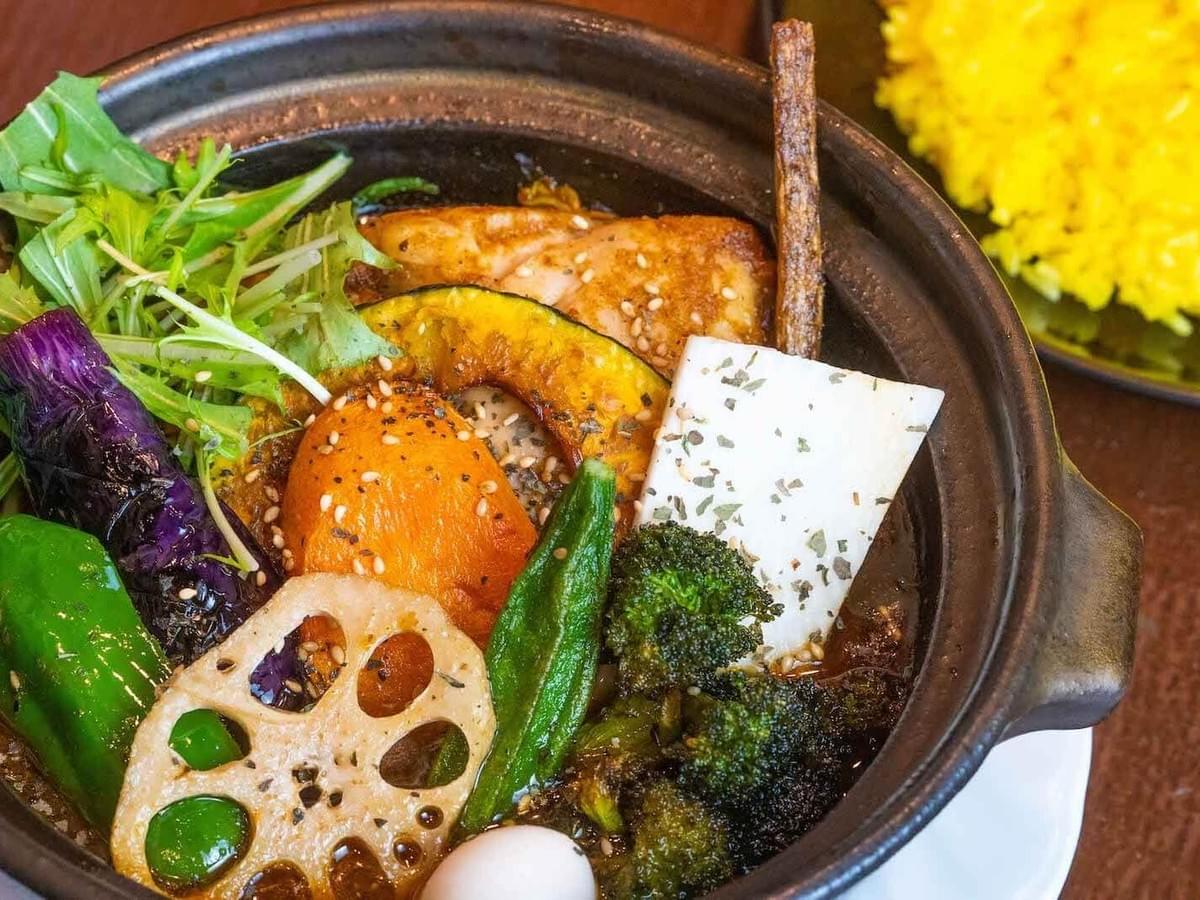 A black ceramic bowl is filled with a colorful Japanese soup curry containing a variety of vegetables such as lotus root, broccoli, pumpkin, okra, and eggplant, garnished with sesame seeds. In the background, a side dish of vibrant yellow turmeric rice is partially visible on a dark plate. A black ceramic bowl is filled with a colorful Japanese soup curry containing a variety of vegetables such as lotus root, broccoli, pumpkin, okra, and eggplant, garnished with sesame seeds. In the background, a side dish of vibrant yellow turmeric rice is partially visible on a dark plate.