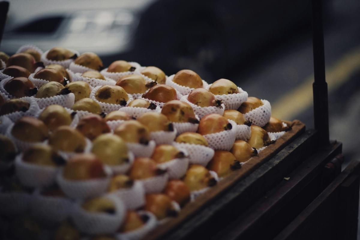 A large cluster of small, pale pomegranates sits nestled in protective white mesh sleeves on a wooden market cart. The image features a shallow depth of field, casting the street background into a moody, dark blur that emphasizes the texture of the fruit. A large cluster of small, pale pomegranates sits nestled in protective white mesh sleeves on a wooden market cart. The image features a shallow depth of field, casting the street background into a moody, dark blur that emphasizes the texture of the fruit.
